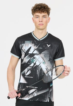 Ung mand i sort sportsbluse med abstrakt mønster holder en badmintonketcher mod en ensfarvet baggrund.