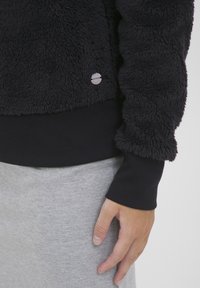 Svart fleecegenser med en strukturert overflate og ribbestrikket mansjett. Har en liten metallisk logo på kanten. Båret over et lysegrå plagg.