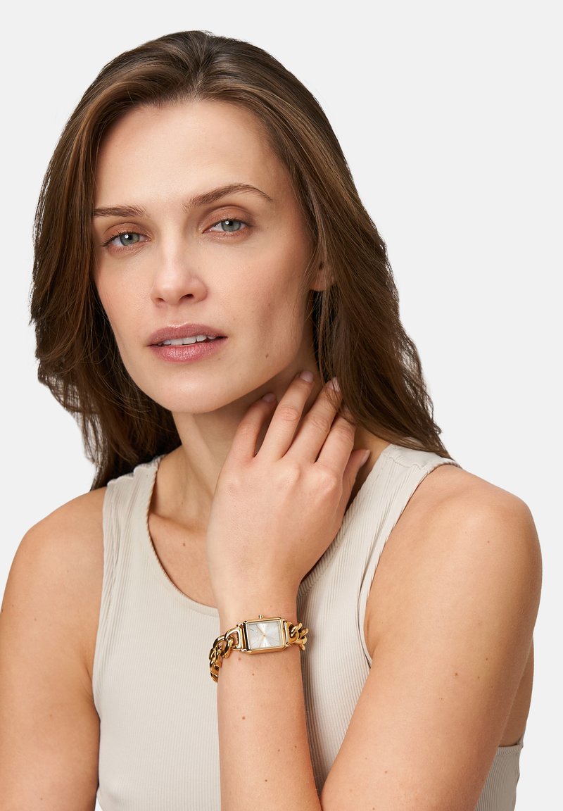 Goldfarbener Armbanduhr mit rechteckigem weißen Ziffernblatt, verknüpftem Kettenarmband und strukturiertem Kronenstück. Auf einem nackten Handgelenk vor einem neutralen Hintergrund getragen.