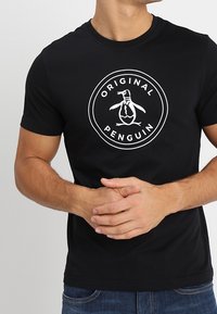 Hombre vestido con una camiseta negra con el logo blanco de "Original Penguin" que presenta un pingüino con corbata, de pie frente a un fondo liso.