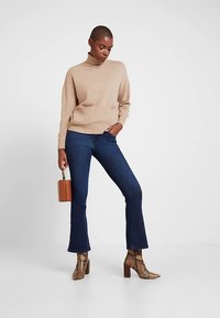 Μπεζ πλεκτό πουλόβερ με ψηλό λαιμό, σε συνδυασμό με σκούρα μπλε τζιν με καμπάνα. Τα παπούτσια έχουν σχέδιο με τύπωμα φιδιού και τακούνια τύπου block.