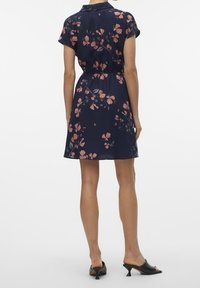 Kvinna klädd i en marinblå kortärmad klänning med rosa blommönster, stående, i kombination med svarta klackskor på en vit bakgrund.