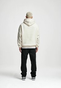 Felpa crema con cappuccio e maniche oversize, abbinata a jeans neri e sneaker bianco e nero. Sfondo neutro.