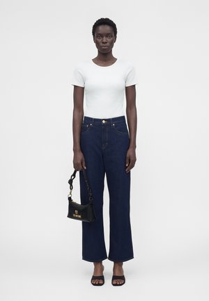 Mujer de pie, vestida con una camiseta blanca de manga corta, vaqueros de talle alto azul oscuro, sandalias negras de tacón y sosteniendo un bolso negro con logo dorado.