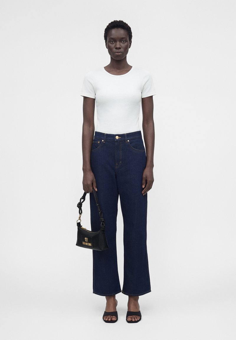 Γυναίκα που στέκεται φορώντας λευκό κοντομάνικο τοπ, σκούρο μπλε τζιν ψηλόμεσο, μαύρα πέδιλα με τακούνι και κρατώντας ένα μαύρο τσαντάκι με χρυσό λογότυπο.