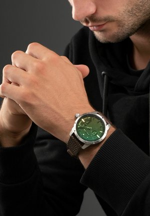 Men's watch with a green patterned dial, silver metal case, and a brown textured leather strap, worn on a hand clasped in front.