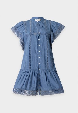 Jeune femme debout les mains dans le dos, portant une courte robe bleue avec des boutons et des détails en dentelle, associée à des chaussures plates noires.
