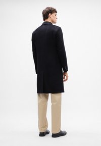 Cappotto lungo nero in lana con colletto a punta, spacchetto singolo sul retro, abbinato a pantaloni beige e scarpe nere. Tessuto morbido, vestibilità su misura.
