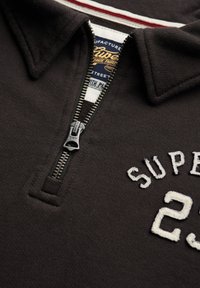 Polo nero con colletto zip, testo ricamato bianco "SUPER" e "23", con una bordatura a righe rosse e bianche sul colletto interno.