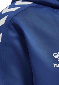 Giacca sportiva blu realizzata in materiale leggero e liscio, con motivi a chevron bianchi lungo le spalle e un logo sul petto.