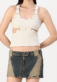 Top corto de color beige con nervaduras y tachuelas, con cortes en los laterales. Combinado con una falda de mezclilla corta en diferentes tonos de azul y marrón claro, con cremallera frontal.