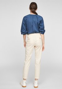 Mujer de pie con la espalda visible, que lleva una blusa azul, pantalones crema con estampado y bolsillos traseros abotonados, además de botas blancas hasta el tobillo.