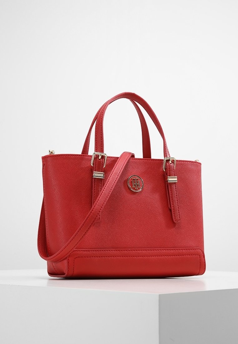 Sac à main en cuir rouge avec deux poignées supérieures, une bandoulière amovible, des boucles dorées et un emblème circulaire du logo à l’avant.