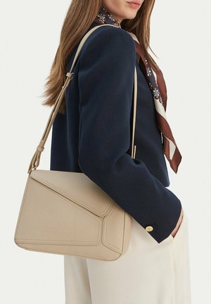 Woman wearing navy blazer and patterned scarf carries beige leather shoulder bag with adjustable strap.