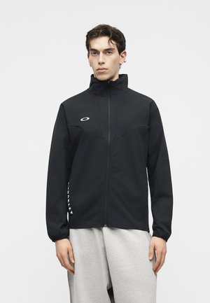 Νεαρός άνδρας με μαύρο μπουφάν με φερμουάρ και ψηλό γιακά και ανοιχτό γκρι παντελόνι, στέκεται μπροστά σε απλό λευκό φόντο.