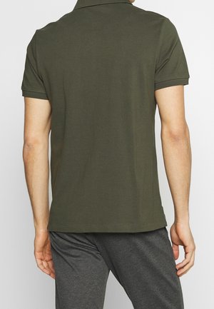 Mand iført en olivengrøn kortærmet poloshirt og mørkegrå bukser, vist bagfra mod en ensfarvet baggrund.