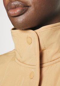 Beige vuorattuja takkeja, joissa on neppari-suljenta, sileä pinta ja huomaamatonta ommellut yksityiskohtia.