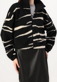 Kobieta w czarno-białej polarowej kurtce w paski zebry, zapiętej na szaro, połączona z czarną skórzaną spódnicą.
