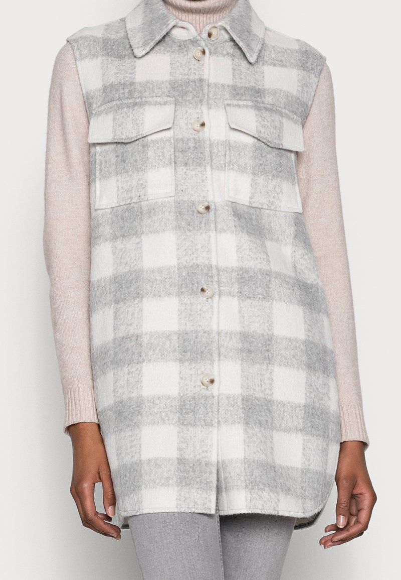 Gilet sans manches à carreaux gris et blanc boutonné, porté par-dessus un pull beige à col roulé et manches longues ainsi qu'un pantalon gris.