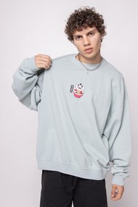 Eine junge Person mit lockigem Haar trägt einen hellblauen Pullover, auf dem ein kleiner gestickter Cartoon eines Pandas mit einer Futterschüssel zu sehen ist.