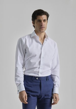 Camisa blanca de manga larga con cuello estructurado, combinada con pantalones ajustados de color azul real con rayas y detalles de botones.