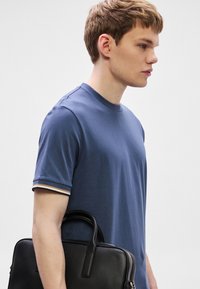 Tengerészkék pamut póló kerek nyakkal és rövid ujjakkal, csíkos díszítéssel az ujjvégeken. Egyik kezében fekete bőr aktatáska.