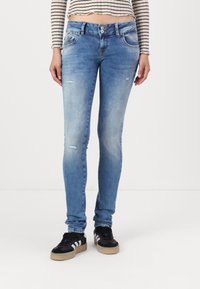 Vaqueros ajustados de color azul claro con detalles desgastados, cierre de botón y cremallera, diseño clásico de cinco bolsillos. Combinados con zapatillas negras.