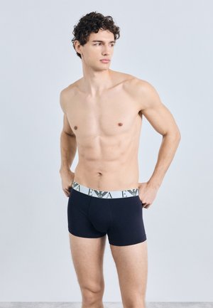 Tumši zili bokseršorti ar baltu jostasvietas gumiju, uz kuras redzams logotips. Materiāls šķiet mīksts, ar pieguļošu dizainu un vidēja garuma līdz augšstilbam.