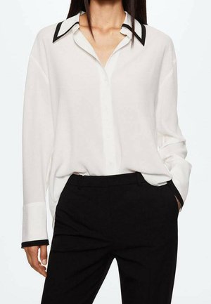 Camisa blanca abotonada con cuello y puños en contraste negro, mangas largas y ajuste holgado, combinada con pantalones negros a medida.