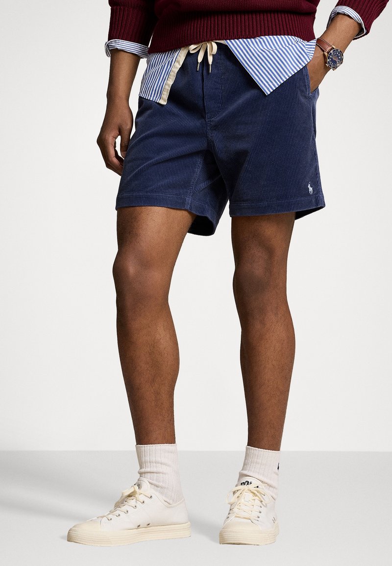 Shorts en velours côtelé bleu marine avec taille élastique à cordon, poches latérales et petit emblème. Associés à des baskets blanches et des chaussettes de couleur claire.