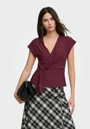 Femme portant un haut cache-cœur sans manches bordeaux et une jupe à carreaux noir et blanc tenant un sac noir sur un fond uni.