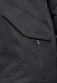 Giacca grigia scura con una grande tasca inclinata e una cerniera verticale. Il materiale sembra liscio con una finitura opaca.