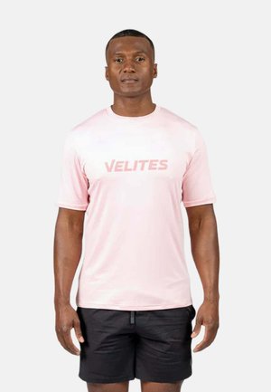 Hombre que lleva una camiseta rosa claro con el texto "VELITES" y pantalones cortos negros, de pie frente a un fondo claro y liso.