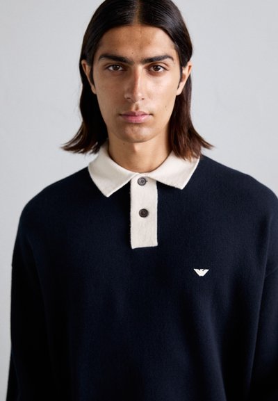 Pulover polo tricotat de navă cu guler de culoare crem și trei nasturi. Logo mic pe piept. Textură moale, croială casual și design minimalist.