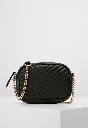 Sac bandoulière en cuir matelassé noir avec bandoulière chaîne dorée et fermeture éclair, présenté sur une surface blanche avec un fond clair uni.