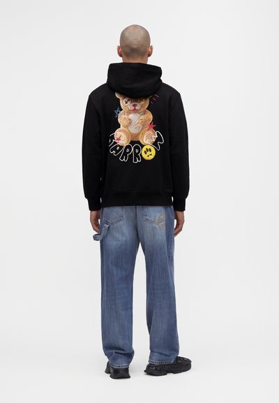 Homme portant un sweat à capuche noir avec un ours en peluche coloré et le texte "NO PROBLEM" dans le dos, associé à un jean bleu ample et des chaussures noires.