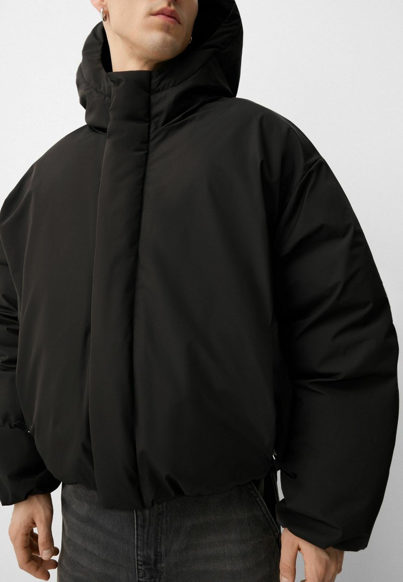 Uomo che indossa una giacca a vento nera imbottita con cappuccio e tasche laterali, insieme a jeans grigio scuro, su uno sfondo chiaro e uniforme.