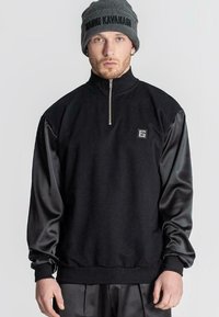 Černý hybridní svetr s límcem na zip, vyrobený z texturované látky a hladkých saténových rukávů. Obsahuje malé označení značky.