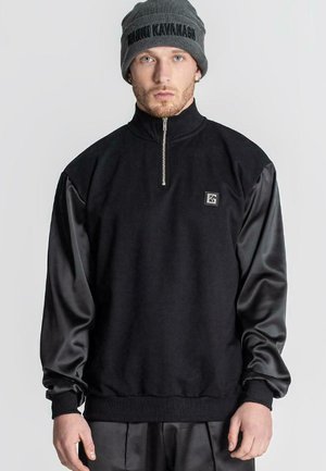 Sudadera híbrida negra con cuello con cremallera, que presenta un tejido texturizado y mangas de satén lisas. Incluye un pequeño logo de la marca.