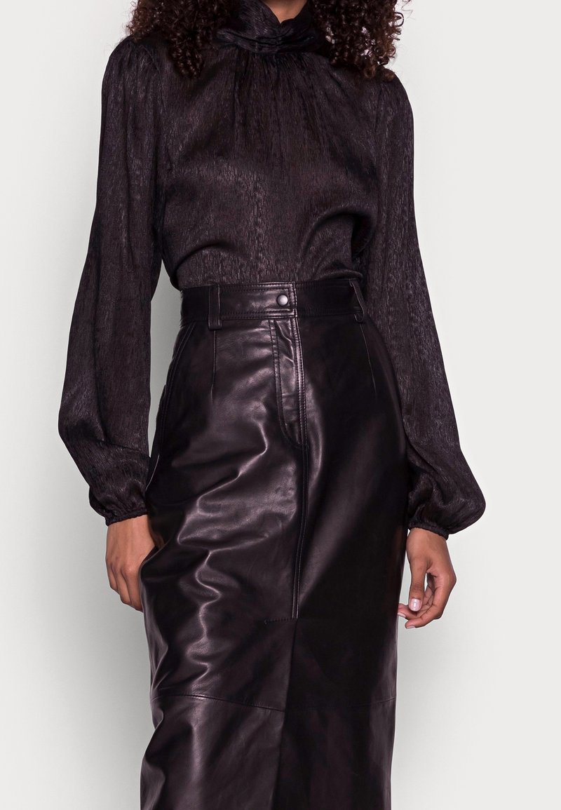 Femme portant un chemisier noir à manches longues avec un col haut et une jupe en cuir noire taille haute sur un fond uni.