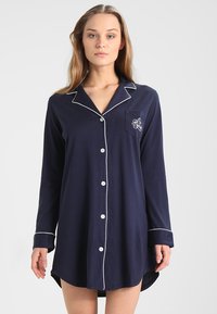 Mørkeblå langermet nattskjorte laget av mykt stoff, med hvite kanter, knappestolpe foran og en brystlomme med broderte detaljer.
