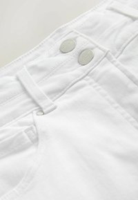 Gros plan sur un jean en denim blanc montrant des boutons doubles argentés et une poche avant avec détail passants de ceinture.