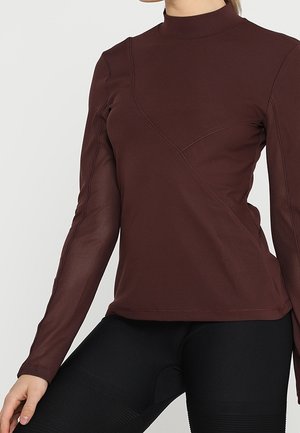 Kvinde iført en tætsiddende brun langærmet top med diagonale sømdetaljer og sorte teksturerede leggings, poserer mod en ensfarvet baggrund.