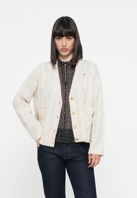 Cardigan beige a maglia con scollo a V, polsini e orlo a costine. Presenta grandi bottoni dorati e tasche frontali a toppa. Indossato sopra una camicetta a pois.