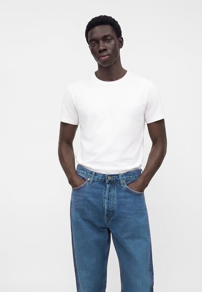 Jeune homme aux cheveux courts et bouclés, portant un t-shirt blanc uni et un jean bleu, debout les mains dans les poches devant un fond uni.