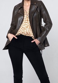 Giacca marrone in ecopelle con cerniera, collo a rever e spalline, abbinata a una camicetta con stampa leopardata e pantaloni neri.