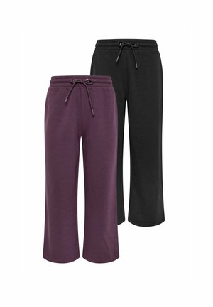 Woman standing against wall wearing a loose purple sweatshirt with shoulder buttons and matching wide-leg pants with white sneakers.