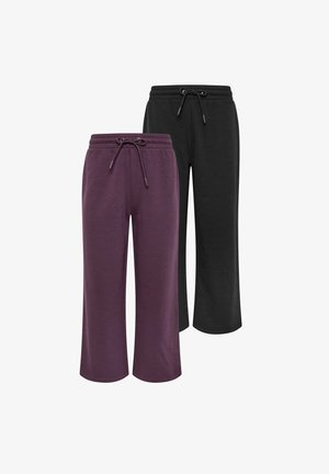 Femme debout contre un mur, portant un sweat-shirt ample violet avec des boutons aux épaules et un pantalon large assorti avec des baskets blanches.