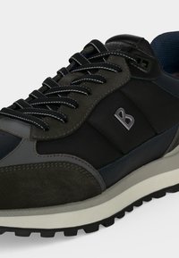 Gros plan d'une basket de couleur foncée avec des détails noirs et gris, une semelle texturée, des lacets réfléchissants et un logo métallique "B" sur le côté.