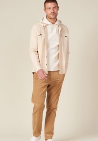 Helles beige Button-Up-Hemd über einem weißen Hoodie, kombiniert mit hellbraunen Cordhosen und weißen Sneakers. Lässige, entspannte Passform mit Taschen.